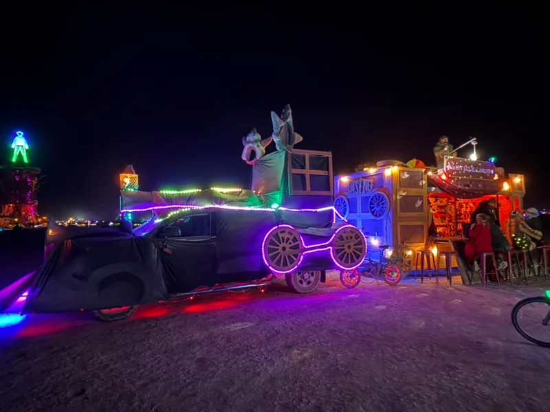 Side view of the art car and wagon lit in purple, with players at the table